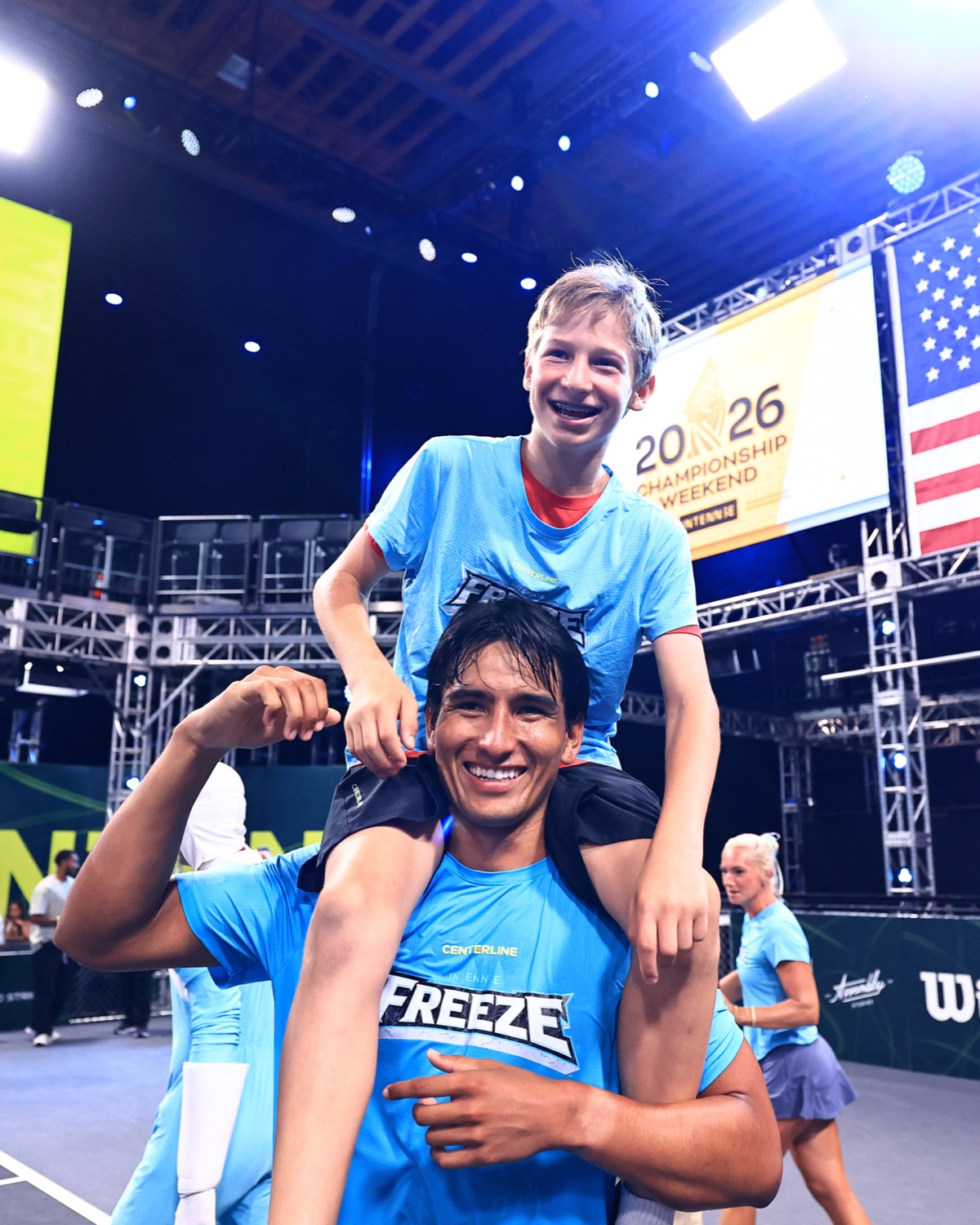 A young ball keeper in Freeze kit riding on the shoulders of a Freeze player, both grinning, under Championship Weekend signage.