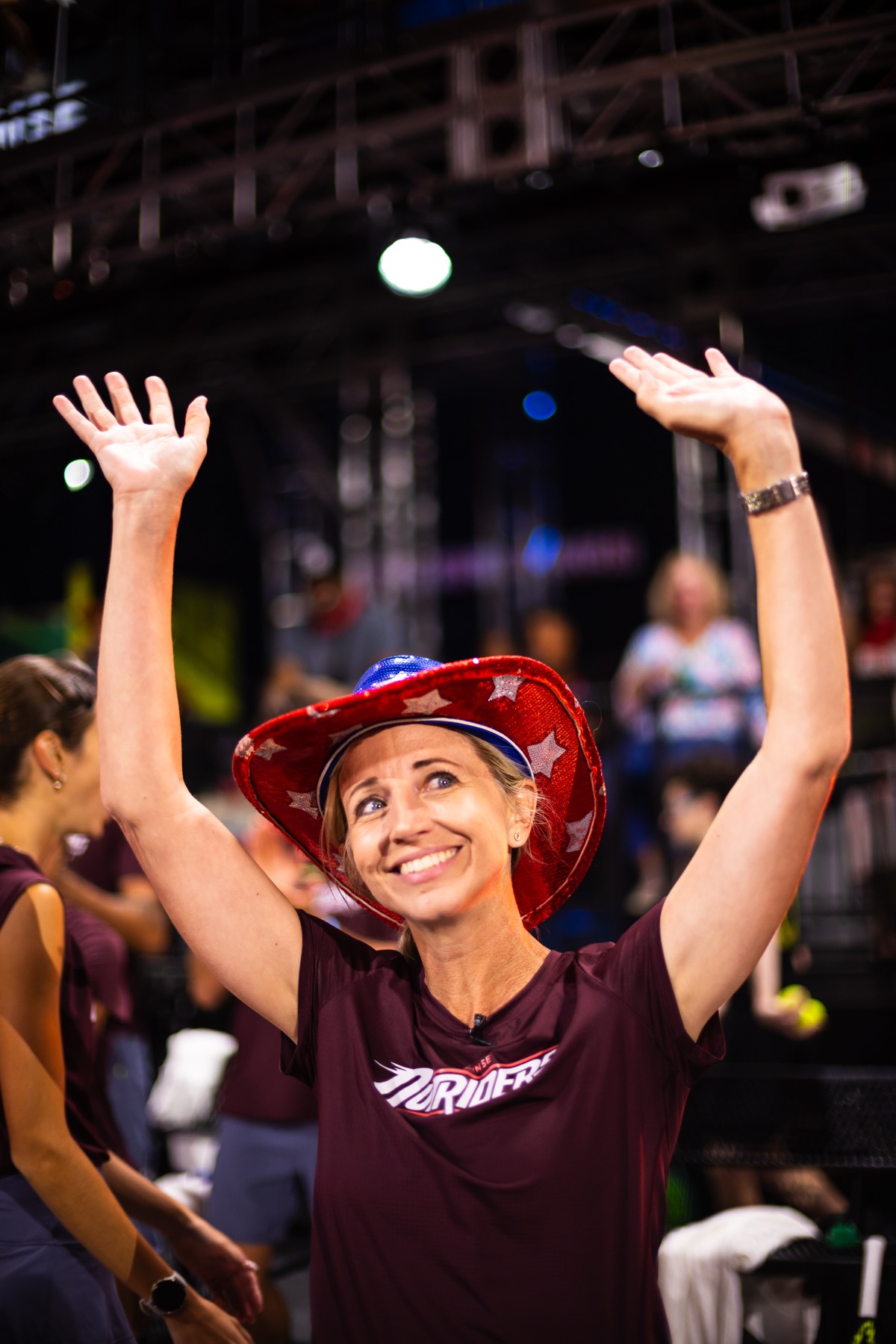 Amy Bartlett O'Connell watching from the stands in a wide-brimmed hat as a player celebrates in front of a full house.
