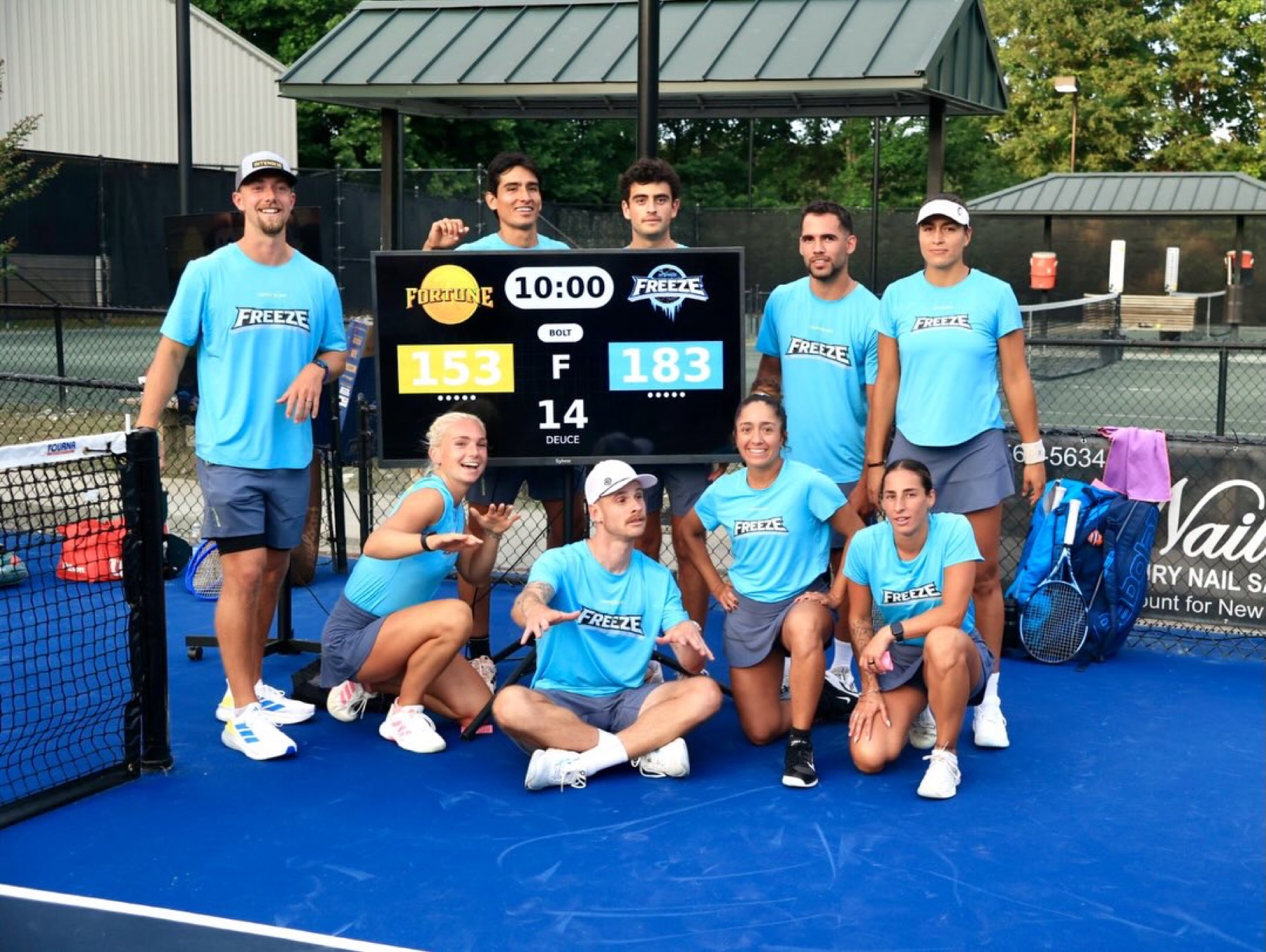 Eight players in matching light blue team kit posed around a portable scoreboard on an outdoor hard court, trees and a pavilion behind.