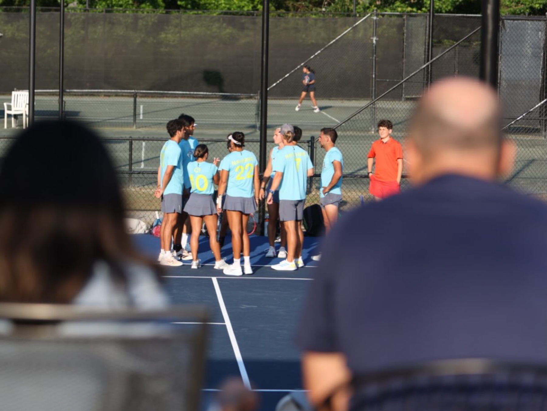 A team huddle on an outdoor court seen over the shoulders of club members who have stopped to watch courtside.