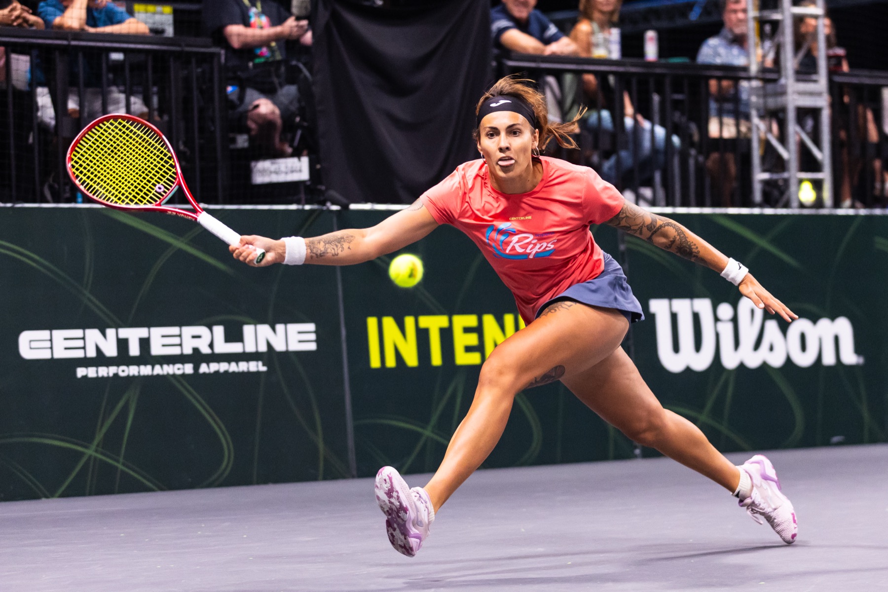 A player stretching wide for a forehand under arena lights, Centerline and Wilson signage behind.